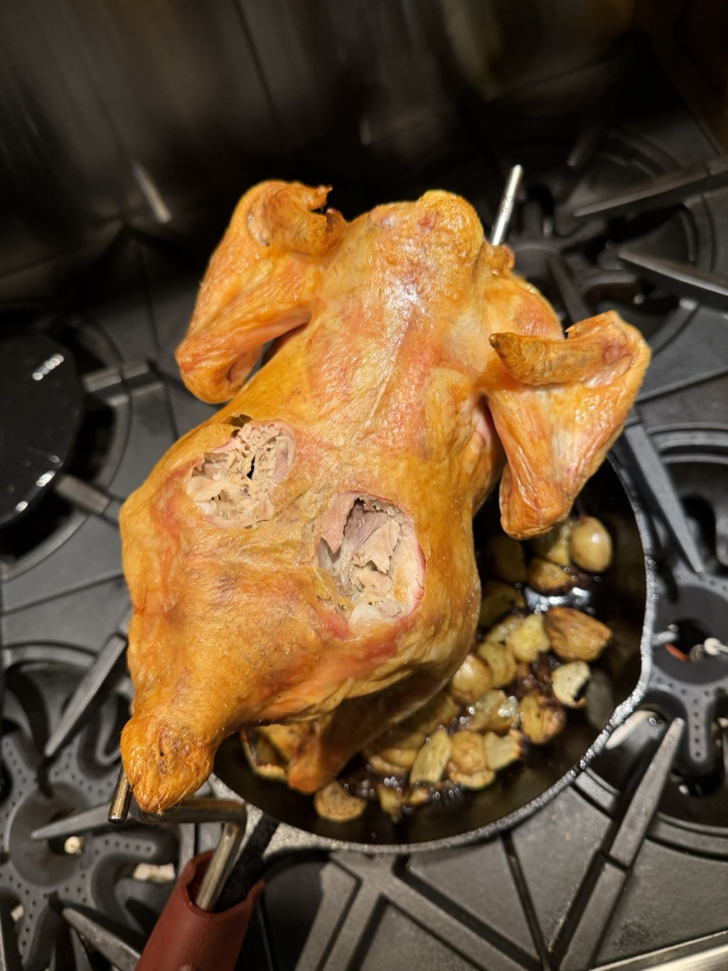 Whole roasted chicken on the PoulTree rod showing the crispy breast side, potatoes in the skillet below