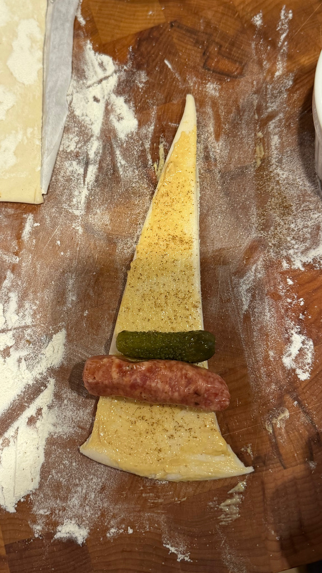 Puff pastry triangle with mustard, celery salt, sausage, and cornichon ready to roll
